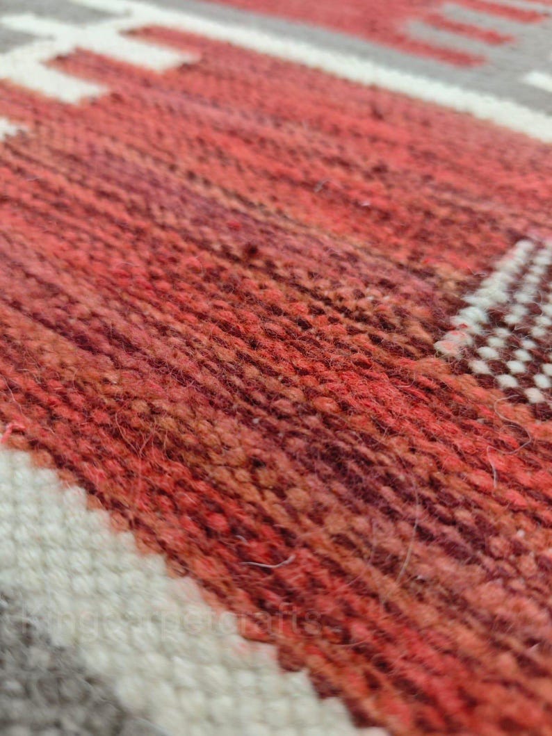 May include: Close-up of a woven rug with a geometric pattern. The rug features a blend of red, orange, and brown hues, with white and gray accents. The texture of the rug is visible, showcasing the woven fibers.