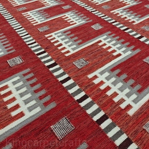 May include: A red and white geometric patterned rug. The rug features a repeating pattern of white, gray, and black shapes against a red background. The design includes rectangular and square elements, creating a textured appearance.