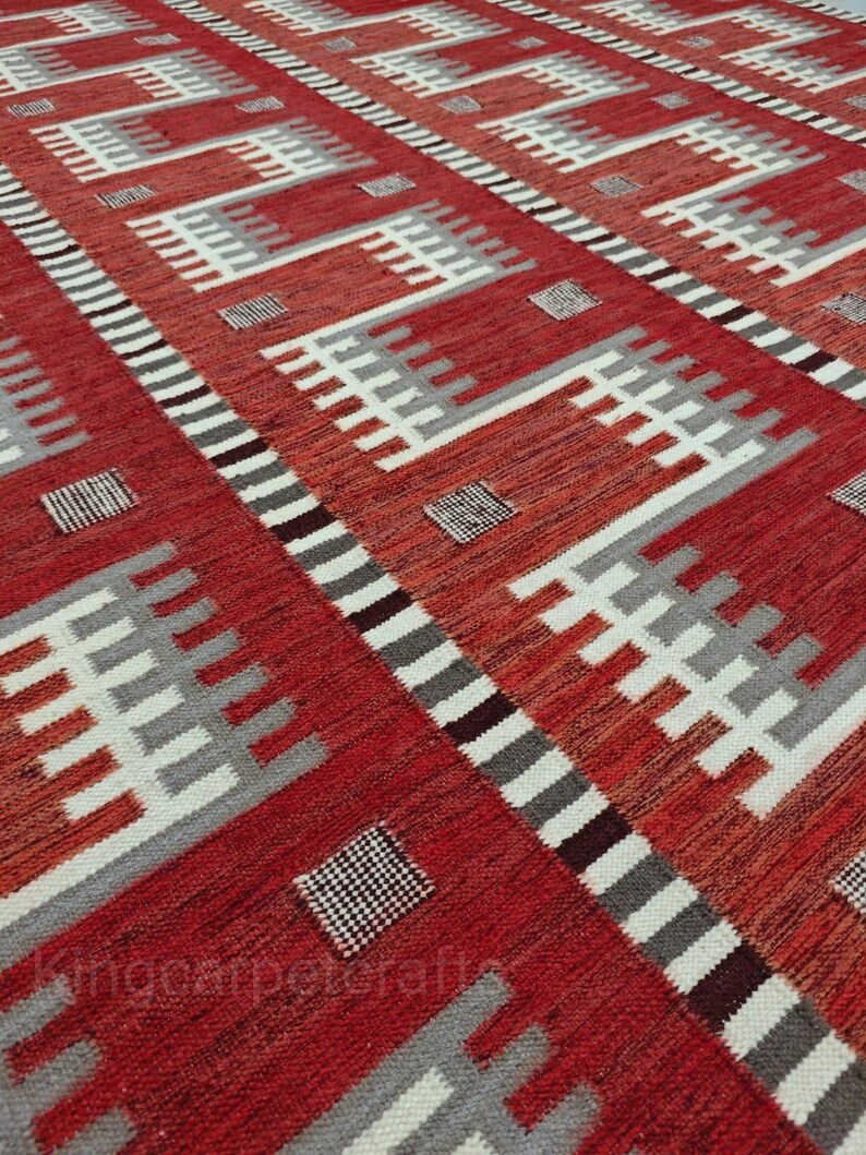 May include: A red and white geometric patterned rug. The rug features a repeating pattern of white, gray, and black shapes against a red background. The design includes rectangular and square elements, creating a textured appearance.