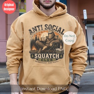 May include: A tan hooded sweatshirt featuring a graphic print of a Sasquatch wearing sunglasses, sitting in a camping chair by a fire. The text reads ANTI SOCIAL SQUATCH CLUB and MEMBERSHIP: ZERO PEOPLE, with a mug labeled LEAVE ME ALONE.
