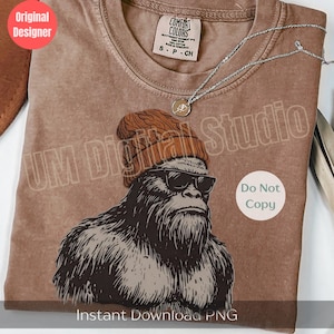 May include: A brown short-sleeve t-shirt featuring a graphic print of a hairy creature wearing a brown knit beanie and black sunglasses. The shirt has a Comfort Colors brand label at the neck and a silver-toned chain necklace resting on top.