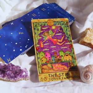 Może przedstawiać: Karta tarota z kolorową ilustracją wróżki, domów i napisem "THE STAR TOOTH FAIRY". Talia niebieskich kart z białymi gwiazdami i półksiężycami znajduje się za kartą tarota. Ametyst, muszla i kamień również na zdjęciu.
