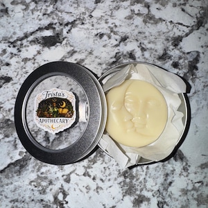 May include: An open, round, silver tin containing a pale yellow, moon-shaped product. The product features a carved design of two faces. The tin also contains a label with the text "Trista's Apothecary".