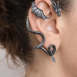 May include: A silver-toned ear cuff featuring a dragon design. The cuff wraps around the ear, with a dragon head at the top and a wing detail. The dragon's body curves down the ear, ending in a curled tail.