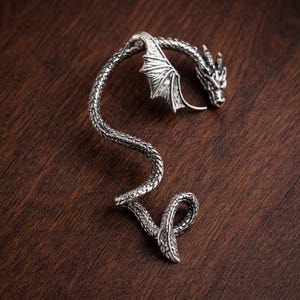 May include: A silver-colored dragon ear cuff. The dragon features detailed scales, wings, and a head with intricate facial features. The cuff curves along the ear, with the dragon's head at the top and the tail ending in a curled design.