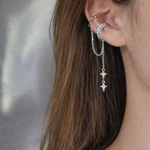 Könnte beinhalten: Ein silberner Ohrklemme mit einer Kette und zwei baumelnden Sternanhängern. Die Ohrklemme umschließt das Ohr und verbindet sich mit einer Kette, die herunterhängt, mit zwei sternförmigen Anhängern am Ende. Der Schmuck ist mit kleinen, funkelnden Steinen besetzt.