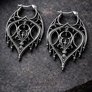 May include: A pair of silver-colored hoop earrings with an ornate, gothic-inspired design. The earrings feature a detailed filigree pattern with a central cross and fleur-de-lis elements, and small dangling beads.