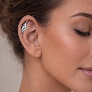 Puede incluir: Un brazalete de oreja plateado con un diseño intrincado se usa en la oreja. El brazalete de oreja se curva alrededor de la oreja externa, con un patrón decorativo en la parte delantera. La joya es un accesorio elegante.