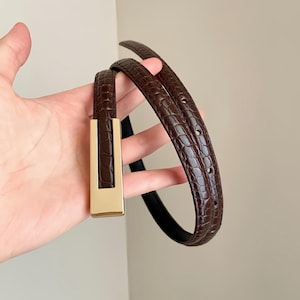 May include: A slim brown belt featuring a textured crocodile-embossed pattern and a rectangular gold-tone metal buckle. The belt strap has a glossy finish and includes several pre-punched holes for adjustment along the length of the band.