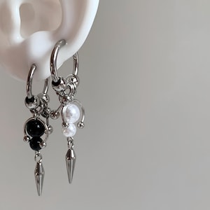 May include: Two silver-toned hoop earrings featuring dangling charms. One earring displays black spherical beads and a silver spike, while the other features white spherical beads and a silver spike. Both hoops are secured with metallic beads.