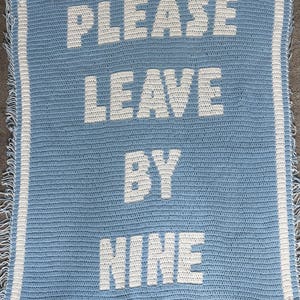 May include: A light blue crocheted blanket with the words "PLEASE LEAVE BY NINE" in white block letters. The blanket has a white border and fringe.