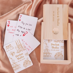 Könnte beinhalten: Eine Holzkiste mit Schiebedeckel, graviert mit "Declan & Thalia" und dem Datum 06.08.2025. Mehrere Spielkarten mit handgeschriebenen Nachrichten und goldenen Illustrationen liegen um die Kiste herum.