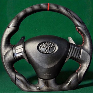May include: A black and carbon fibre Toyota steering wheel with red stitching and a red stripe at the top. The wheel has integrated controls and paddle shifters. The Toyota logo is in the centre.