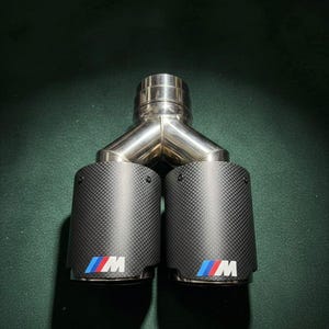 Könnte beinhalten: Ein Doppel-Auspuffsystem mit Carbonfaser-Endrohren. Der Auspuff hat ein poliertes Y-Rohr aus Edelstahl und zwei zylindrische Endrohre mit dem BMW M-Logo in Blau, Rot und Weiß. Die Endrohre haben ein Carbonfaser-Webmuster.