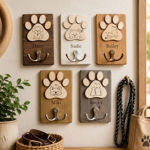Könnte beinhalten: Fünf rechteckige Wandhaken aus Holz, jeweils mit einem hellen Holz-Pfotenabdruck, einer eingravierten Tierillustration und einem Namen: Daisy, Sadie, Bailey, Milo und Rocky. Jedes Schild ist mit einem doppelten Metallhaken versehen.