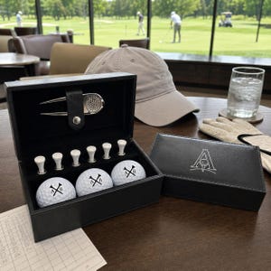 Könnte beinhalten: Ein Golf-Geschenkset aus schwarzem Kunstleder mit drei weißen Golfbällen mit A- und B-Logo, sechs weißen Tees und einer silbernen Pitchgabel in einer ausgekleideten Box. Der Deckel zeigt ein weißes A-Logo mit Golfballgrafik.