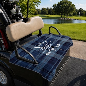 Puede incluir: Una funda de asiento para carrito de golf a cuadros azules y negros con el nombre Oliver en texto blanco, acompañada de iconos gráficos blancos de palos de golf, una pelota de golf y tres estrellas, sobre el asiento de un carrito negro.