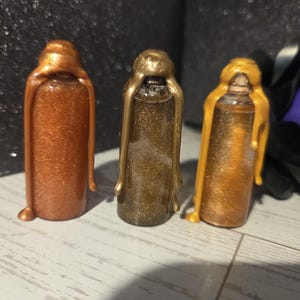May include: Three small, glitter-filled bottles with metallic wax-like seals. The bottles are in shades of copper, bronze, and gold. The wax appears to be dripping down the sides of the bottles. The background is a dark, glittery surface.