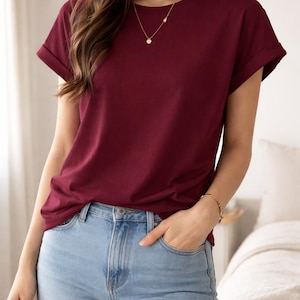 May include: A burgundy t-shirt with rolled-up sleeves and a crew neck. The shirt is paired with light blue denim jeans. A gold necklace and bracelet are visible.