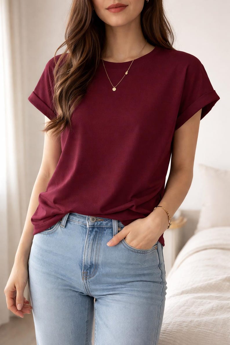 May include: A burgundy t-shirt with rolled-up sleeves and a crew neck. The shirt is paired with light blue denim jeans. A gold necklace and bracelet are visible.