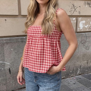 Può includere: Un top senza maniche a quadri rossi e bianchi con scollo quadrato e spalline annodate. Il top è abbinato a jeans blu. Il top ha una vita arricciata e una vestibilità ampia.