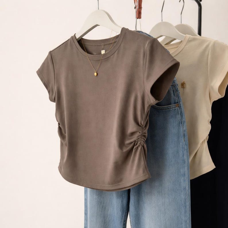 May include: A taupe-colored t-shirt with short sleeves and a gathered side detail. The shirt hangs on a white hanger with a gold pendant necklace. Light blue jeans are visible below the shirt.