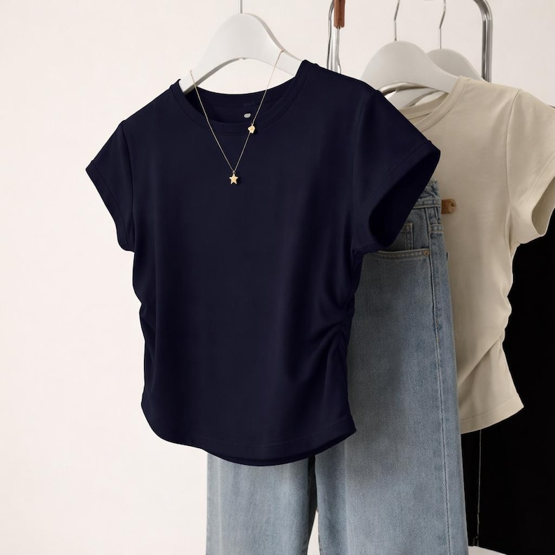 May include: A navy blue short-sleeved t-shirt with a gathered side detail, hanging on a white hanger. A gold star pendant necklace is visible. Light wash denim jeans are also in the image, along with a beige t-shirt.