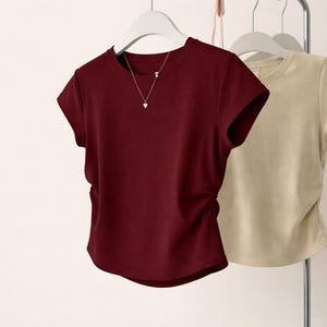 May include: Two short-sleeved t-shirts hanging on a rack. One is a deep burgundy color, and the other is a light beige. The burgundy shirt has a gold necklace with a heart pendant. Both shirts have a crew neck.