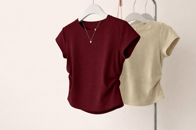 May include: Two short-sleeved t-shirts hanging on a rack. One is a deep burgundy color, and the other is a light beige. The burgundy shirt has a gold necklace with a heart pendant. Both shirts have a crew neck.