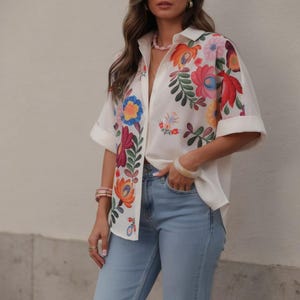 Puede incluir: Camisa blanca con bordado floral colorido. La camisa tiene mangas cortas y dobladas y se combina con vaqueros azul claro. El diseño floral incluye flores rojas, naranjas, rosas y azules.