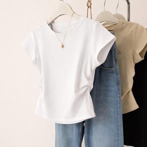 May include: A white short-sleeved t-shirt with a round neckline hangs on a hanger, paired with blue jeans. A gold-colored necklace with a pendant is visible. Other shirts in beige and black are also on hangers.