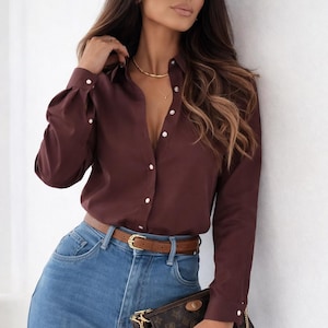 May include: A burgundy shirt with long sleeves and white buttons, worn with blue jeans and a brown belt. A small brown purse with a gold chain is also visible.