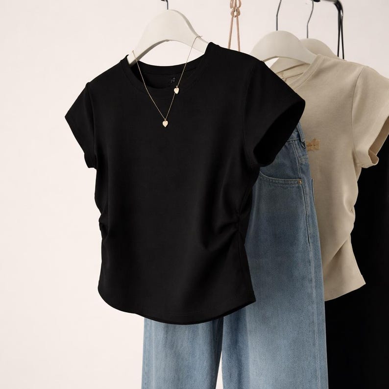 May include: A black short-sleeved t-shirt with a gathered side detail, hanging on a white hanger. A gold necklace with heart pendants is visible. Light blue denim jeans are also in the image, along with a tan t-shirt.