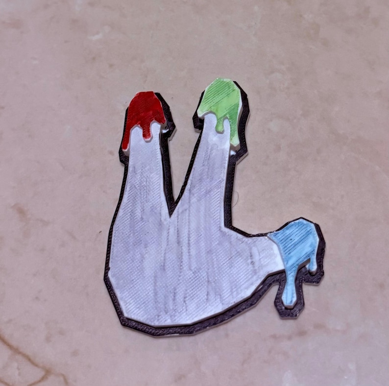 May include: A white, abstract-shaped object with a black outline, featuring red, green, and blue paint drips. The design has a textured appearance, suggesting a 3D-printed or crafted item. The overall aesthetic is modern and artistic.