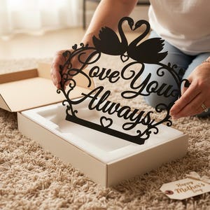 Könnte beinhalten: Schwarzes Metallschild mit den Worten "Love You Always" und zwei Schwanensilhouetten. Das Schild wird in einer weißen Box mit einem beigefarbenen Kartondeckel gehalten. Ein Etikett mit "Happy Mother's Day" ist sichtbar.