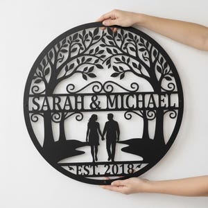 Könnte beinhalten: Ein kreisförmiges Wandbild aus schwarzem Metall mit einem lasergeschnittenen Motiv eines Baumes mit ausladenden Ästen. In der Mitte steht der Text SARAH & MICHAEL mit einer Silhouette zweier Händchen haltender Personen und EST. 2018 unten.