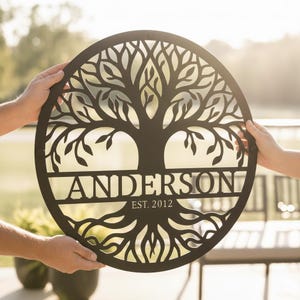 Könnte beinhalten: Ein rundes Wandbild aus schwarzem Metall mit der Silhouette eines Baumes, dessen Äste nach oben und Wurzeln nach unten ragen. Ein horizontaler Balken in der Mitte zeigt den Namen ANDERSON mit dem darunter stehenden Text EST. 2012.