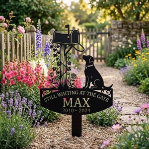 Könnte beinhalten: Schwarzer Metall-Gartenstecker mit Briefkasten, Hundesilhouette und der Aufschrift "Still Waiting at the Gate", dem Namen "MAX" und den Daten 2010-2024. Der Stecker ist von bunten Blumen umgeben, ein Gedenkgarten-Thema.