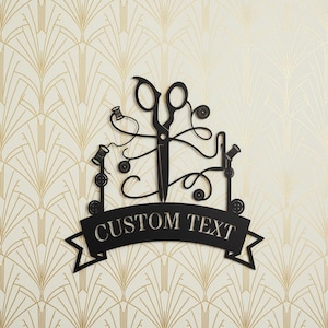 Könnte beinhalten: Schwarze Metall-Wandkunst mit Schere, Garnrollen und Knöpfen. Das Design beinhaltet ein Banner mit dem Text "CUSTOM TEXT". Der Hintergrund hat ein gold-cremefarbenes Art-Deco-Muster.