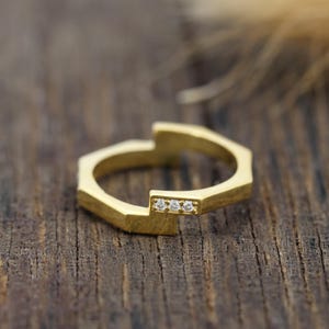 May include: A gold ring featuring an open, geometric, hexagonal-inspired band design. One side of the band is accented with three small, clear, round-cut stones set in a horizontal row, creating a subtle sparkle against the polished gold metal.