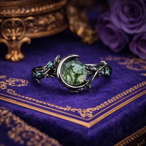 May include: A silver ring with a crescent moon holding a green moss sphere. The ring has detailed vine and leaf patterns, with small blue-green stones. The ring is set on a purple velvet surface.