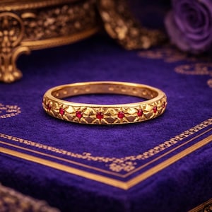Peut inclure: Une bague dorée ornée d'un motif répétitif en forme d'étoiles et de petites pierres précieuses rouges. La bague repose sur une surface de velours violet profond avec des bordures dorées et des motifs floraux. Un bijou luxueux.