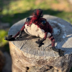 May include: A handmade, crocheted frog with a mohawk. The frog features a red and black color scheme on its head and legs, with a cream-colored body. It has large, black eyes and is posed on a wooden stump.