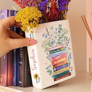 May include: A white ceramic vase shaped like a book, decorated with floral illustrations and the names of classic novels. The vase is filled with colorful dried flowers. The name "Sophia" is written on the side.