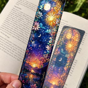 May include: A decorative bookmark with a stained-glass design featuring a celestial theme. The bookmark showcases a vibrant sunset over a lake, a sun, moon, and floral elements. The name "Sophia" is printed on the bookmark.