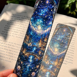 May include: A decorative bookmark featuring a stained-glass design with a night sky, castle, and bridge scene. The bookmark has a dark border and is held over an open book. The color palette includes blues, golds, and whites.
