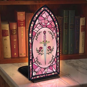 Könnte beinhalten: Eine rosa Buntglaslampe in Bogenform mit einem zentralen ovalen Design mit einem Schwert, Rosen und Blättern. Die Lampe hat einen schwarzen Sockel und ist beleuchtet, mit einem Bücherregal im Hintergrund.