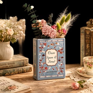 May include: A blue ceramic vase in the shape of a book, featuring the text "Once Upon a Time" and floral designs. The vase contains a bouquet of pink and green flowers. Other books and a teacup are visible in the background.