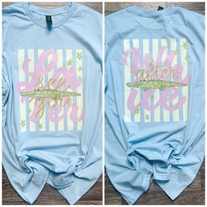 May include: Light blue long-sleeve shirt with a design on the front and back. The front features the text "See You" in pink script, a green alligator, and gold stars. The back says "Wild After Ice" with the same alligator and stars.