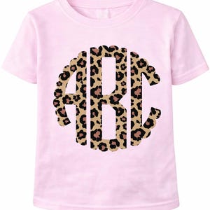 May include: A light pink short-sleeved t-shirt featuring a large, circular monogram graphic in the center. The monogram consists of the letters ABC in a scalloped-edge circle, all filled with a brown and black leopard print pattern.
